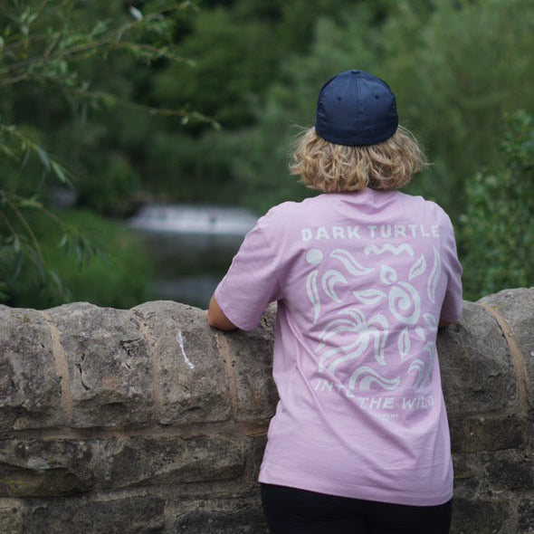 Person wearing a pink t-shirt with a graphic design, standing by a stone wall with greenery in the background.