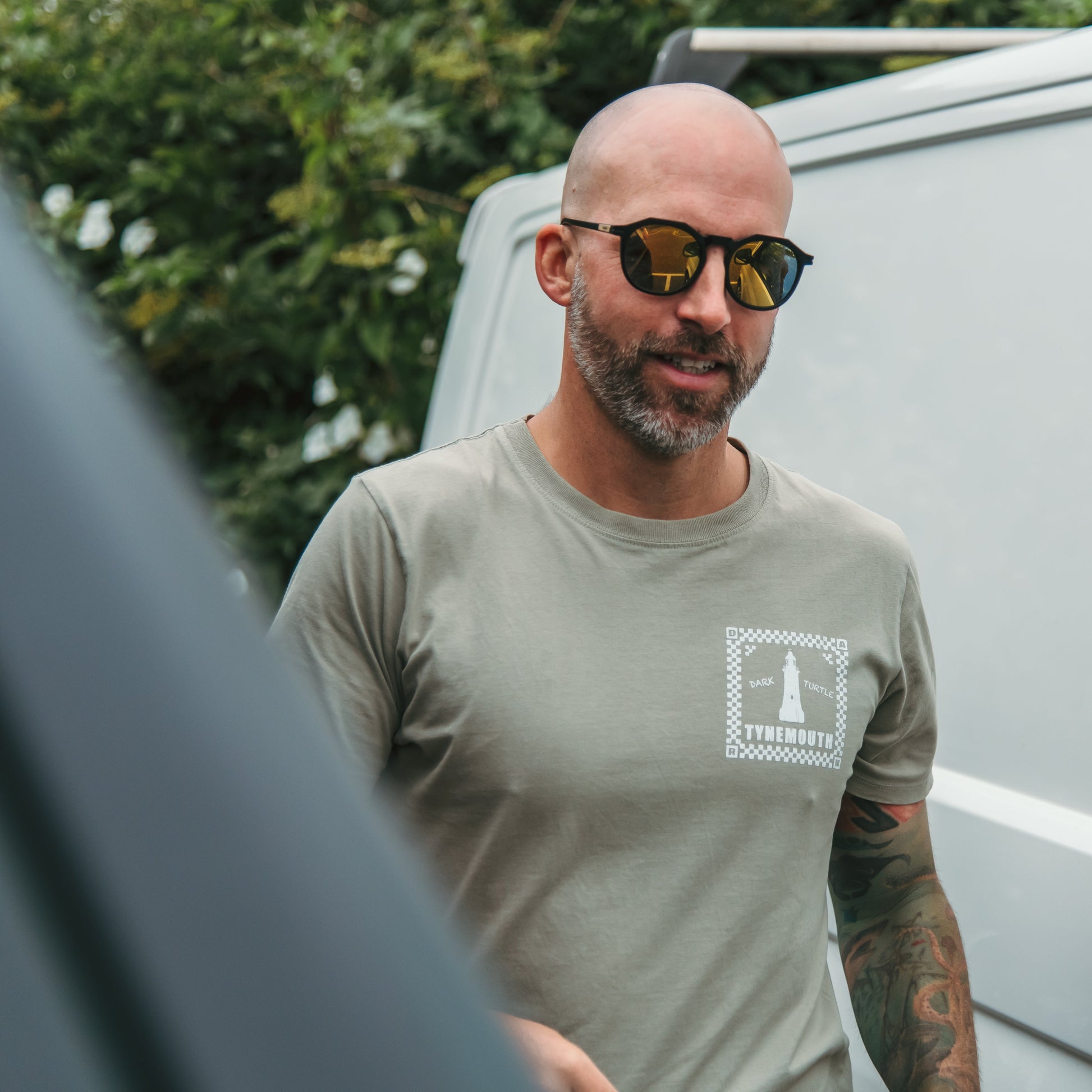 Man wearing sunglasses and a green t-shirt with a logo, standing next to a white van.