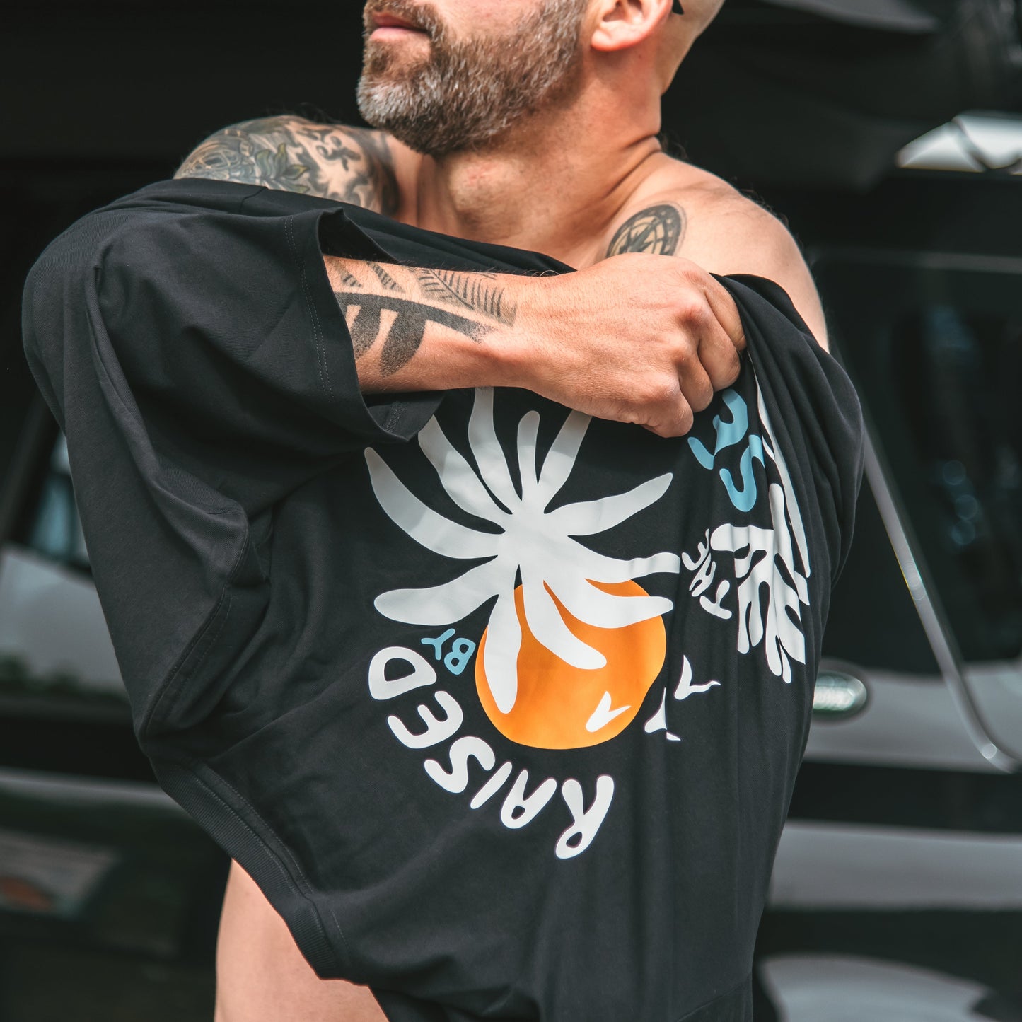 Man wearing a black t-shirt with a graphic design, standing in front of a vehicle.