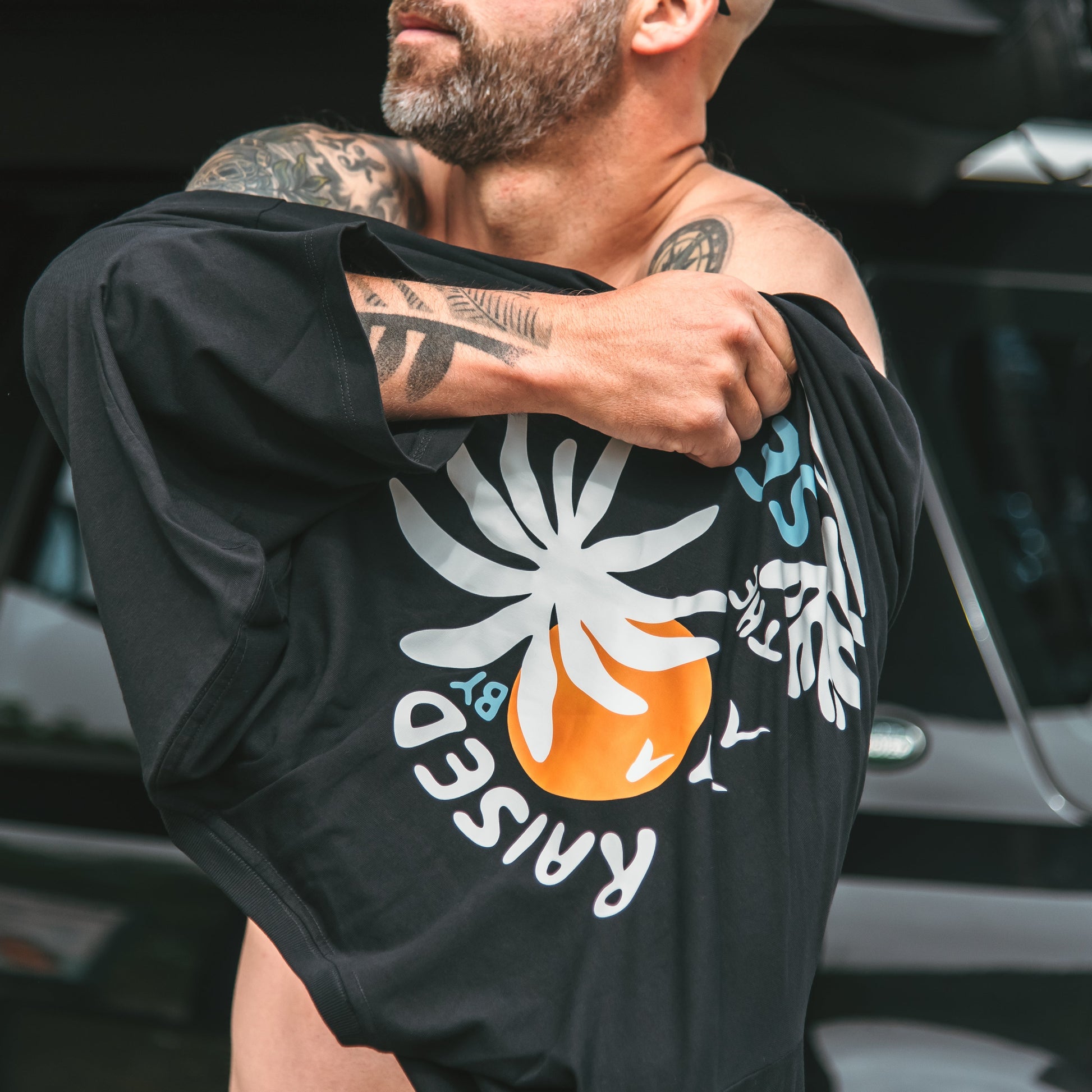 Man wearing a black t-shirt with a graphic design, standing in front of a vehicle.